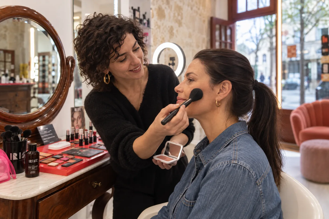 Pendant une séance pour apprendre à se maquiller à Paris, la maquilleuse montre le geste du blush à une participante, devant le mur en pierre de la boutique du Quartier latin.